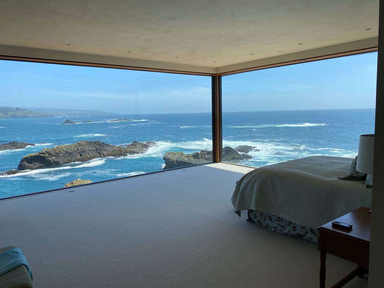 Cliffside bedroom — corner glass walls, turquoise ocean, waves on lava rock